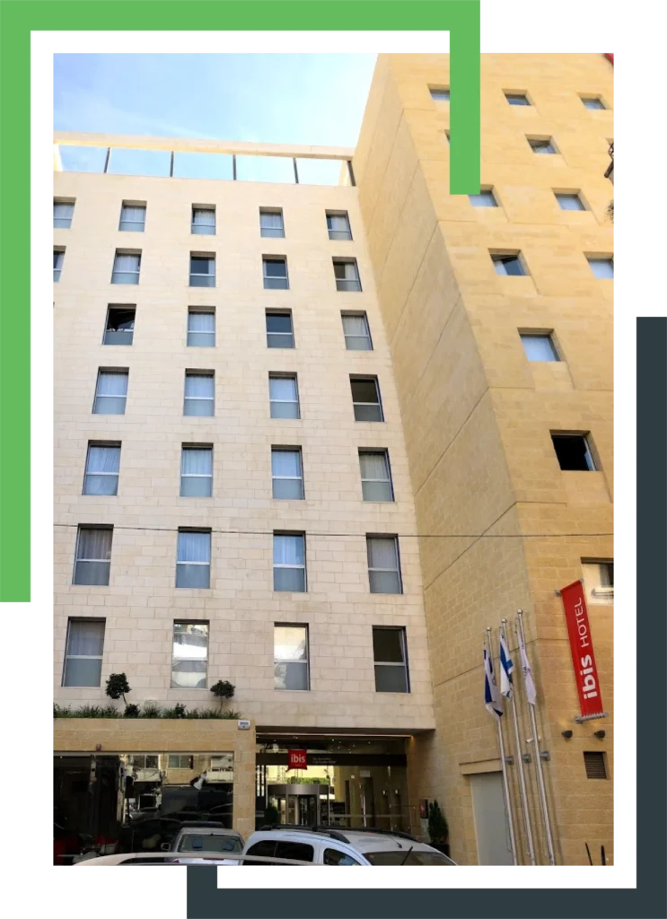 בניין מלון בגוון בהיר עם חלונות מלבניים, דגלים בכניסה ושלט אדום עם הכיתוב "ibis hotel."